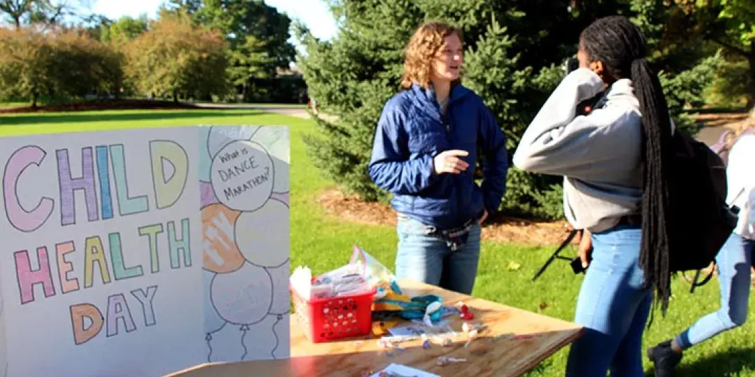 Two young people talk outside next to a table with a Child Health Day sign on it.