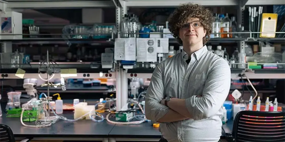 A college student with arms crossed stands in a science lab with equipment all around him.