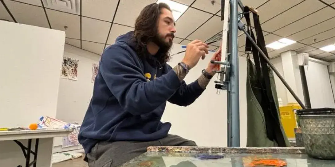 A male college student with long brown hair and a goatee paints on a canvas.