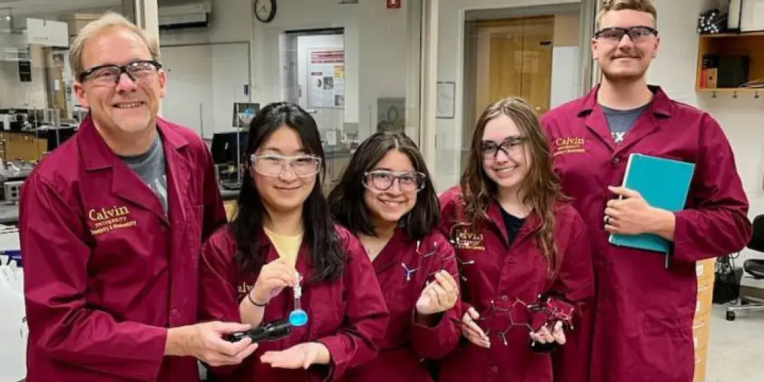 A professor and four students all in maroon lab coats smile at the camera.