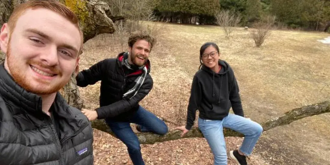 Three students in a photo sitting on a long branch with a field behind them.