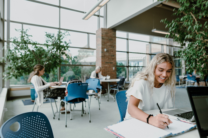 A female student writes in a notebook inside the DeVos Communications building, with tables, chairs and trees surrounding.