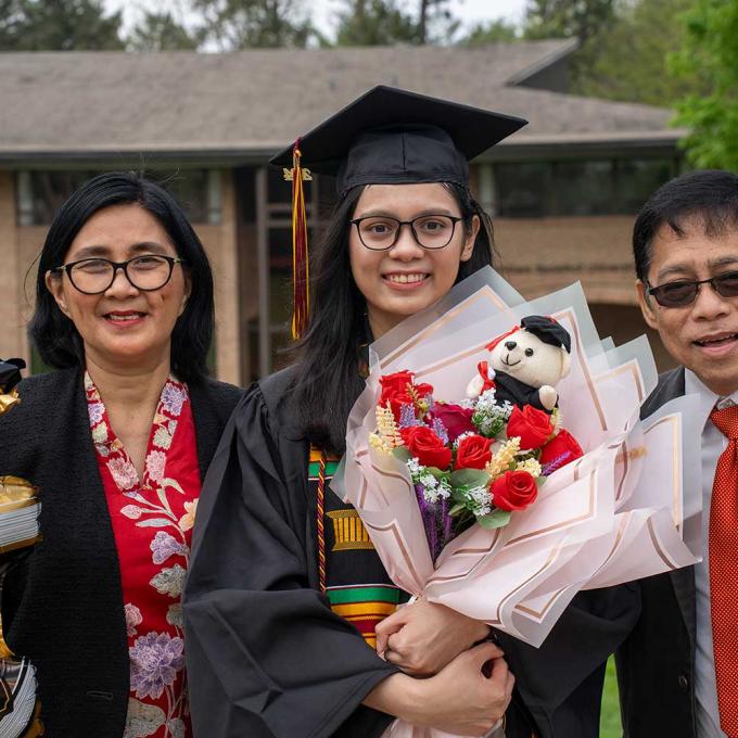 Two Calvin parents standing with their graduate in between who is holding flowers