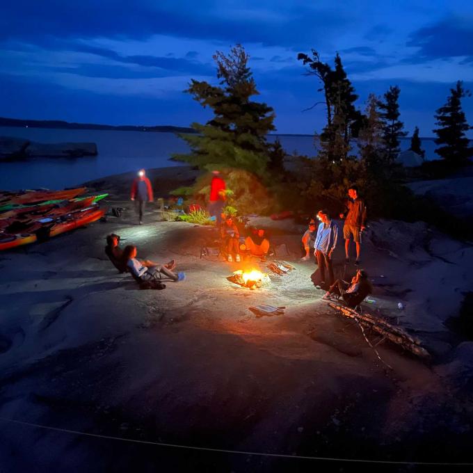 A group of students circles a campfire at dusk, wearing headlamps, with kayaks on the lakeshore behind them.