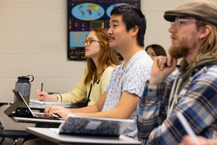 Three students sit in a classroom looking toward the front, with notebooks and laptops open.