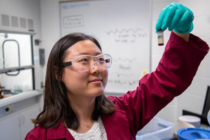 A student holds a lab sample in a tube while wearing a maroon lab coat and safety glasses.