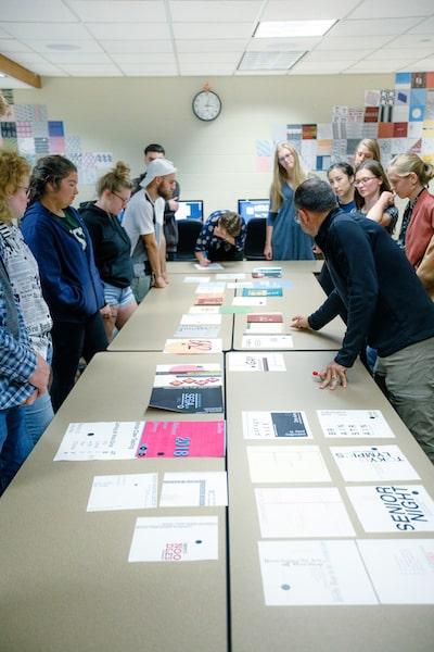 A group of students and a professor stand around tables while evaluating printed designs.