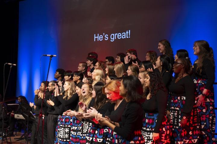 Gospel choir sings and claps, wearing black tops and colorful skirts, with a slide that reads, He's great! in the background.