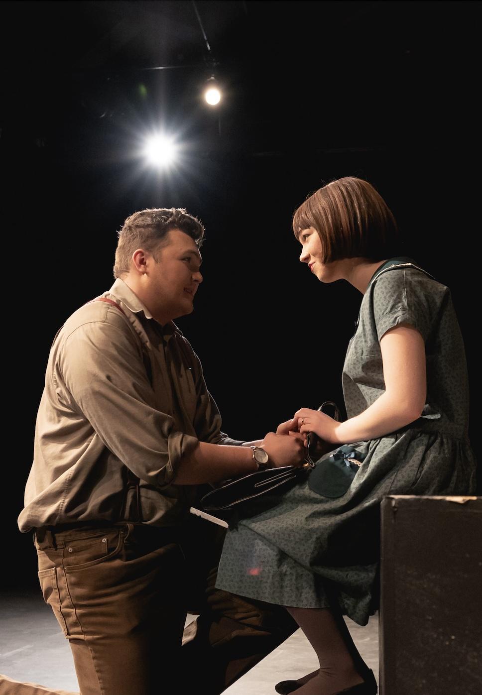 Two actors on stage, looking into each others faces. A theater light shines in the background.